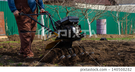 Man cultivating rich soil with a walk-behind tractor in a backyard garden, tilling ground for spring planting on a small farm homestead, preparing beds for growing food. Man cultivating rich soil with a walk-behind tractor in a backyard garden, tilling ground for spring planting on a small farm homestead, preparing beds for growing food. 140508282