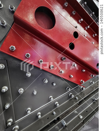Close view of a machine part showing red and black sections with bolts and holes in an industrial setting Close view of a machine part showing red and black sections with bolts and holes in an industrial setting 140508621