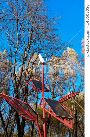 Small wind turbine and solar panels installed on a red metal structure in a park. Alternative renewable energy source against a blue sky and bare trees. Small wind turbine and solar panels installed on a red metal structure in a park. Alternative renewable energy source against a blue sky and bare trees. 140509050