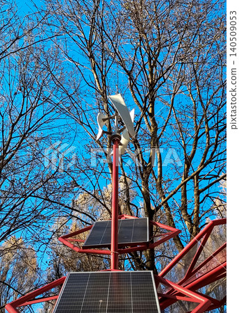 Small wind turbine and solar panels installed on a red metal structure in a park. Alternative renewable energy source against a blue sky and bare trees. Small wind turbine and solar panels installed on a red metal structure in a park. Alternative renewable energy source against a blue sky and bare trees. 140509053