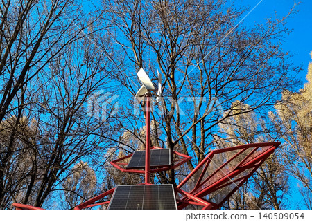 Small wind turbine and solar panels installed on a red metal structure in a park. Alternative renewable energy source against a blue sky and bare trees. Small wind turbine and solar panels installed on a red metal structure in a park. Alternative renewable energy source against a blue sky and bare trees. 140509054