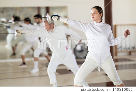 Men and women in white uniforms practice fencing techniques with foils in hands Men and women in white uniforms practice fencing techniques with foils in hands 140510095