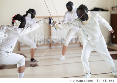 Fencing training - men and women in white uniforms practice fencing in a gymnasium for duel 140510363