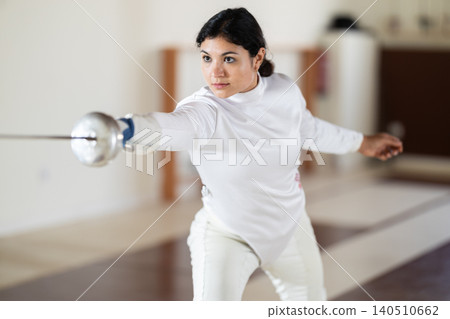 Female athlete training attack movements with rapier at fencing workout in gym Female athlete training attack movements with rapier at fencing workout in gym 140510662