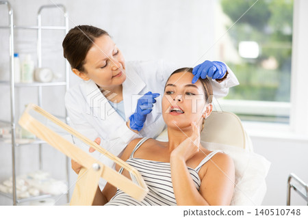 Woman cosmetologist examines patients forehead after lifting procedure and palpates forehead area Woman cosmetologist examines patients forehead after lifting procedure and palpates forehead area 140510748