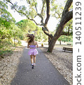 Woman runs on pathway through a park surrounded by trees during a sunny day 140511239