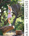 Young woman jumps on a log in a green forest during bright daylight while wearing a pink outfit and sneakers 140511265
