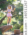 Young woman balances on a tree stump in a sunny forest during daytime while wearing a light outfit 140511267
