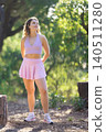 Young woman stands in a park with trees and sunlight while wearing a pink outfit and sneakers 140511280