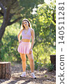 Young woman stands in forest area during daytime while wearing sports attire and posing near tree stump 140511281