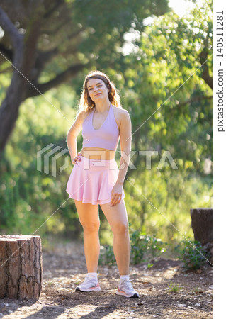Young woman stands in forest area during daytime while wearing sports attire and posing near tree stump Young woman stands in forest area during daytime while wearing sports attire and posing near tree stump 140511281