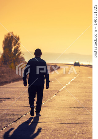 A man walks along a concrete road along the sea at a golden sunset and looks at the mountain A man walks along a concrete road along the sea at a golden sunset and looks at the mountain 140511325