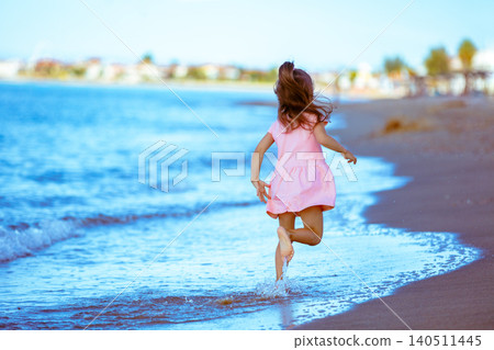 Happy little girl in a pink dress runs along the beach back to the camera in summer Happy little girl in a pink dress runs along the beach back to the camera in summer 140511445