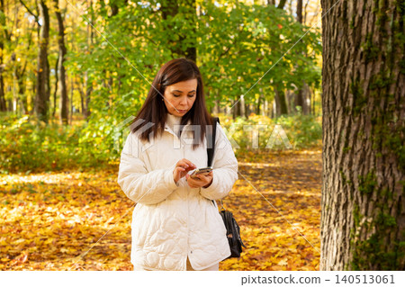 Woman using smartphone in autumn park. Girl texting on mobile device while walking. Casual lifestyle concept, outdoor fall season communication, connection to internet in nature scenery. Woman using smartphone in autumn park. Girl texting on mobile device while walking. Casual lifestyle concept, outdoor fall season communication, connection to internet in nature scenery. 140513061