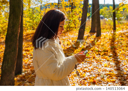 Woman standing in autumn park using smartphone. Girl holding mobile device to check message outdoors. Technology in nature during fall season. Leisure time and digital communication concept. Woman standing in autumn park using smartphone. Girl holding mobile device to check message outdoors. Technology in nature during fall season. Leisure time and digital communication concept. 140513064