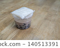 Thai dessert of black beans in sweet coconut milk served in a plastic cup with a side of sticky rice in a clear bag. Placed on a light brown wooden table in a professional stock photography style. 140513391
