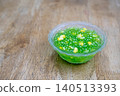 Thai traditional dessert of green tapioca pearls with sweet corn served in a clear bowl on a wooden table. A colorful and authentic Southeast Asian street food snack. Sweet sago pudding with corn. 140513393