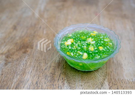 Thai traditional dessert of green tapioca pearls with sweet corn served in a clear bowl on a wooden table. A colorful and authentic Southeast Asian street food snack. Sweet sago pudding with corn. Thai traditional dessert of green tapioca pearls with sweet corn served in a clear bowl on a wooden table. A colorful and authentic Southeast Asian street food snack. Sweet sago pudding with corn. 140513393