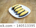 Five roasted bananas in dark skins served on a white plate over a wooden background. A popular healthy snack or dessert in various cultures. High angle view showing the texture of the fruit. 140513395