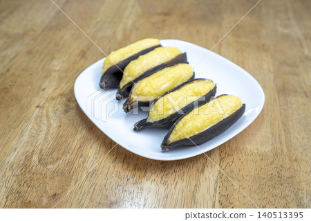 Five roasted bananas in dark skins served on a white plate over a wooden background. A popular healthy snack or dessert in various cultures. High angle view showing the texture of the fruit. Five roasted bananas in dark skins served on a white plate over a wooden background. A popular healthy snack or dessert in various cultures. High angle view showing the texture of the fruit. 140513395