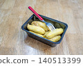 Steamed baby bananas in a black plastic container with a red metal fork on a wooden table. Traditional healthy snack or tropical fruit dessert. High angle view of nutritious street food. 140513399