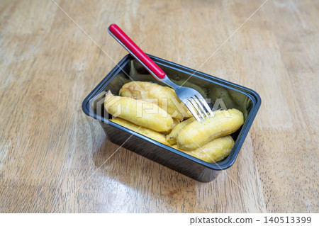Steamed baby bananas in a black plastic container with a red metal fork on a wooden table. Traditional healthy snack or tropical fruit dessert. High angle view of nutritious street food. Steamed baby bananas in a black plastic container with a red metal fork on a wooden table. Traditional healthy snack or tropical fruit dessert. High angle view of nutritious street food. 140513399