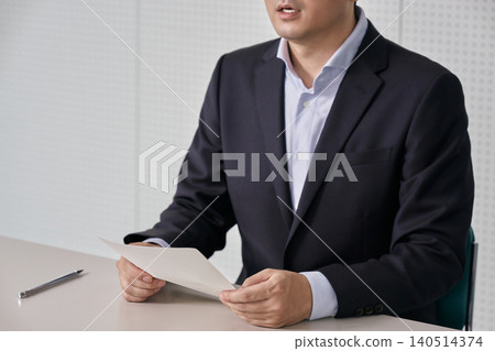 一張沒有臉孔的照片,照片中一名身穿西裝的男子坐在會議桌旁,手持文件,在記者會上發表聲明或演講。 一張沒有臉孔的照片,照片中一名身穿西裝的男子坐在會議桌旁,手持文件,在記者會上發表聲明或演講。 140514374