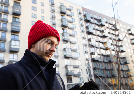 Man in red beanie using mobile phone outdoors near apartment building. Urban guy looking at smartphone screen on city street. Casual lifestyle and communication in cold winter weather. Man in red beanie using mobile phone outdoors near apartment building. Urban guy looking at smartphone screen on city street. Casual lifestyle and communication in cold winter weather. 140514561