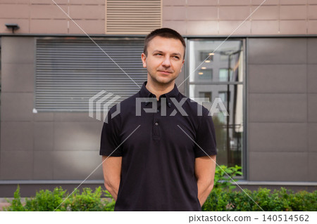 A man stands outside a gray building during daylight. He has a serious expression and wears a dark shirt. Green plants surround him, adding some color to the setting. A man stands outside a gray building during daylight. He has a serious expression and wears a dark shirt. Green plants surround him, adding some color to the setting. 140514562