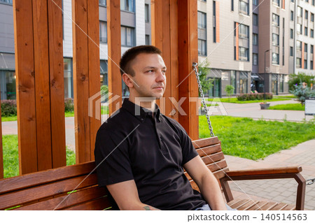 Young man sitting on a wooden hanging bench in a modern residential urban courtyard. Relaxed guy resting outdoors in a stylish housing complex. City lifestyle and residential comfort. Young man sitting on a wooden hanging bench in a modern residential urban courtyard. Relaxed guy resting outdoors in a stylish housing complex. City lifestyle and residential comfort. 140514563