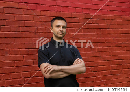 Man standing with arms crossed against a red brick wall. Casual portrait of a confident guy in a black polo shirt. Masculine lifestyle, urban outdoor street photography session. Man standing with arms crossed against a red brick wall. Casual portrait of a confident guy in a black polo shirt. Masculine lifestyle, urban outdoor street photography session. 140514564