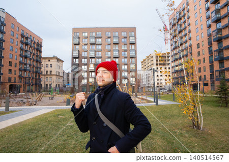Real estate agent standing in front of new residential building complex. Professional man in red hat looking at housing project. Property development, construction and investment concept. Real estate agent standing in front of new residential building complex. Professional man in red hat looking at housing project. Property development, construction and investment concept. 140514567