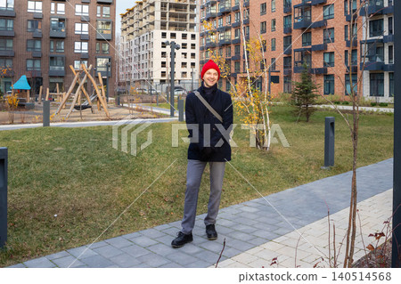 Man standing in a modern residential courtyard. Smiling young guy wearing a red hat and coat walking near apartment buildings in autumn. Urban lifestyle and neighborhood recreation area. 140514568