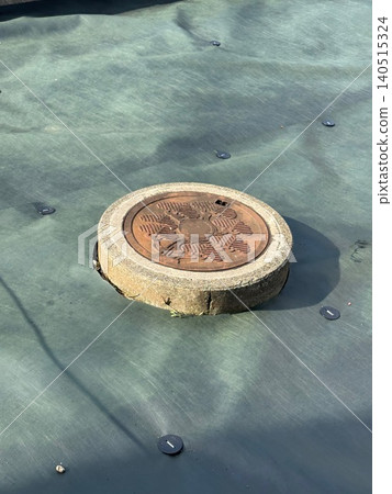 A rusty, round manhole cover exposed from beneath a weed-proof sheet. A rusty, round manhole cover exposed from beneath a weed-proof sheet. 140515324