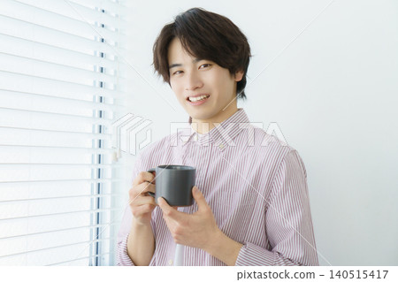 一幅年輕日本男子站在窗邊的肖像,他穿著條紋襯衫,手裡拿著馬克杯。 一幅年輕日本男子站在窗邊的肖像,他穿著條紋襯衫,手裡拿著馬克杯。 140515417