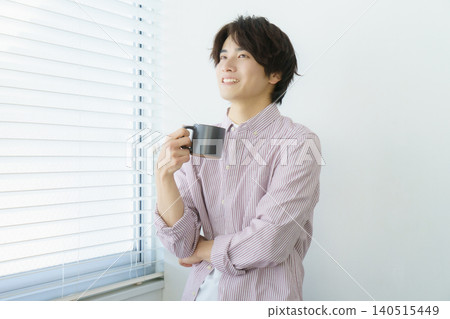 一幅年輕日本男子站在窗邊的肖像,他穿著條紋襯衫,手裡拿著馬克杯。 一幅年輕日本男子站在窗邊的肖像,他穿著條紋襯衫,手裡拿著馬克杯。 140515449