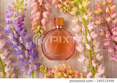 Glass bottle of luxury perfume surrounded by colorful lupine flowers on wooden surface. Fragrance concept for beauty and skincare. Elegant cosmetic product display for spring holiday. 140515496