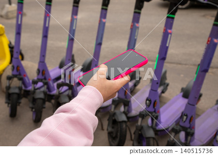 A woman uses a smartphone to activate or end electric scooter rental via a mobile app. Rental urban transport service concept. Modern technology for eco friendly city commute, shared mobility device 140515736