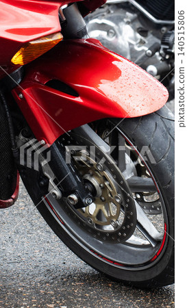 Close up detail shot of red motorcycle front wheel, fender, tire, and brake disc with water droplets on wet asphalt road, vertical photo composition. Close up detail shot of red motorcycle front wheel, fender, tire, and brake disc with water droplets on wet asphalt road, vertical photo composition. 140515806