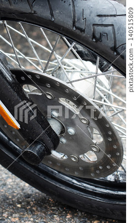 Close-up detail of front wheel of motorcycle on wet asphalt, showcasing tire tread, spokes, disc brake, and suspension fork with orange reflector, day outdoor, vertical photo Close-up detail of front wheel of motorcycle on wet asphalt, showcasing tire tread, spokes, disc brake, and suspension fork with orange reflector, day outdoor, vertical photo 140515809