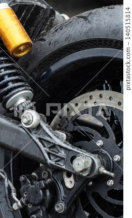 Close up vertical shot of motorcycle rear wheel assembly featuring wet black tire, gold shock absorber, black suspension spring, silver brake disc, and brake caliper component detail, vertical photo 140515814