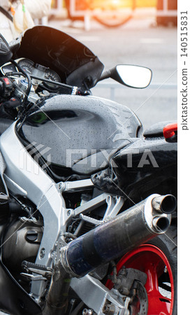 Black sport motorcycle with water droplets on sleek body, silver frame highlighting design, chrome exhaust pipes, vibrant red rear wheel, detail view of parked vehicle after rain or wash, vertical Black sport motorcycle with water droplets on sleek body, silver frame highlighting design, chrome exhaust pipes, vibrant red rear wheel, detail view of parked vehicle after rain or wash, vertical 140515831