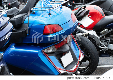 Blue touring motorcycle rear section detail showing chrome luggage rack and multiple taillights, surface covered with water droplets from rainfall, another red motorcycle background. Blue touring motorcycle rear section detail showing chrome luggage rack and multiple taillights, surface covered with water droplets from rainfall, another red motorcycle background. 140515923