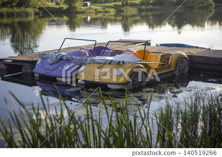 Vintage-style pedal boats shaped like cars moored at a wooden pier on a calm lake Vintage-style pedal boats shaped like cars moored at a wooden pier on a calm lake 140519266