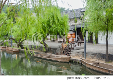 一艘木製觀光船停泊在倉敷川的河面上,柳樹在倉敷地區(岡山縣倉敷市)的秀麗景色中搖曳。 一艘木製觀光船停泊在倉敷川的河面上,柳樹在倉敷地區(岡山縣倉敷市)的秀麗景色中搖曳。 140519520