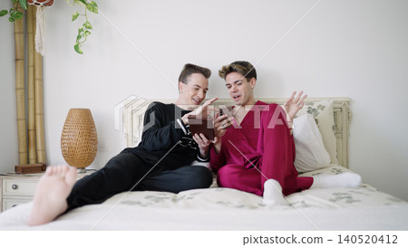 Gay couple relaxing in pajamas on bed using smartphone Gay couple relaxing in pajamas on bed using smartphone 140520412