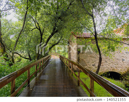 Wet wooden walkway leading past a traditional stone mill building under a canopy of lush green trees along the Uima River trails in Lobao Portugal Wet wooden walkway leading past a traditional stone mill building under a canopy of lush green trees along the Uima River trails in Lobao Portugal 140521355