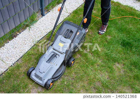 Lawn mower with power cord stands on backyard grass near fence and gravel border Lawn mower with power cord stands on backyard grass near fence and gravel border 140521368