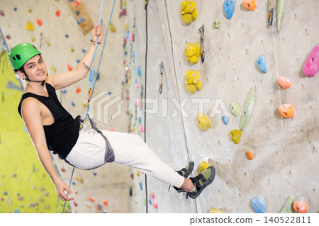 Sporty cheerful young male climber in helmet using safety belts and helmet going down after training in bouldering gym 140522811