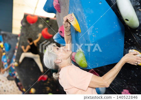 Sporty elderly female exercising in boulder climbing hall without rope, reaching new results, enjoying new challenges in amusement park 140522846